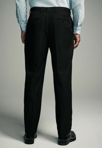 Schwarze Hose mit einem taillierten Schnitt, flacher Vorderseite und zwei Gesäßtaschen. Getragen mit einem hellblauen Langarmhemd und schwarzen Schuhen.