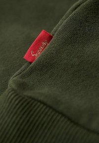 Grön sweatshirt-tyg med en mjuk textur, med en röd varumärkesetikett sydd med guldtext på kanten.