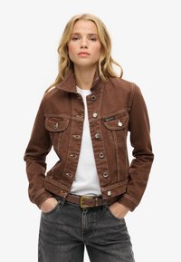Veste en denim marron avec un col haut, une fermeture à boutons, deux poches poitrine et des coutures contrastées. Portée par-dessus une chemise blanche et un jean gris.