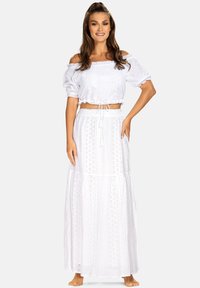 Biały top z odkrytymi ramionami i krótkim krojem, z bufiastymi rękawami i koronkowymi detalami, w zestawieniu z długą, warstwową białą spódnicą z wzorami w ozdobne otwory.