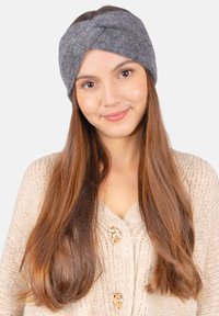 Bandeau en tricot gris avec un design torsadé, fabriqué en laine douce. Porté par une femme aux longs cheveux bruns et lisses, sur un fond neutre.