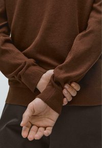 Hnědý pletený svetr s žebrovanými manžetami, s měkkou texturou a přiléhavým střihem. Ruce sepnuté za zády, ukazující uvolněnou postavu.