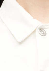 Άσπρο ενδύμα με γιακά και μεταλλικό κουμπί με χαραγμένο το «Boss» κοντά στο λαιμό, σε ανοιχτόχρωμο δέρμα.