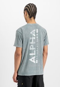 Mees, kellel on punutud juuksed, kannab kulunud sinakas-roheline Alpha Industries T-särk ja musti lühikesi pükse, seistes valge tausta poole seljaga.