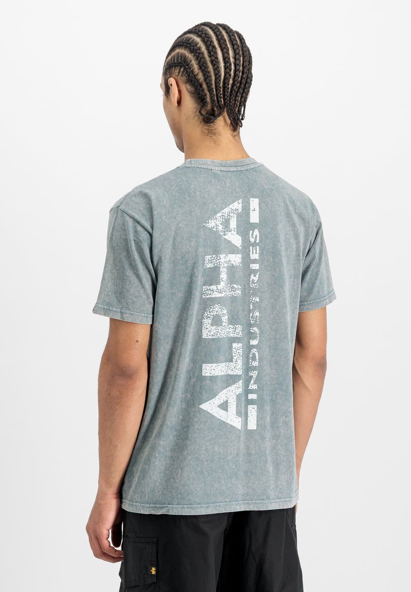 Mees, kellel on punutud juuksed, kannab kulunud sinakas-roheline Alpha Industries T-särk ja musti lühikesi pükse, seistes valge tausta poole seljaga.