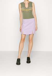 Žena v olivově zeleném grafickém bezramínkovém topu, lila mini sukni s předními rozparky a černých nazouvacích botách na jednobarevném pozadí.