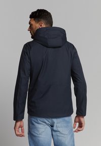 Navyblauwe hoodie jack gemaakt van een gladde stof, met een relaxte pasvorm en een naaddetail aan de achterkant, gecombineerd met lichtblauwe jeans.