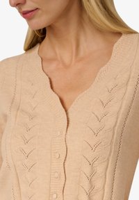 Jasno brzoskwiniowy kardigan z dekoltem w serek, skomplikowanym wzorem liści, żebrowanymi krawędziami oraz zapięciem na guziki z przodu. Miękka dzianinowa struktura.