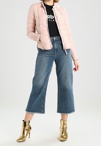 Světle růžová péřovka, černé tričko s grafikou, zkrácené modré džíny s roztrhaným lemem a lesklé zlaté kotníčkové boty.