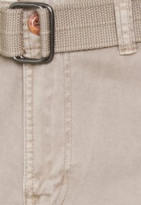 Pantalones de algodón beige con un cinturón trenzado, que cuenta con una hebilla metálica, costuras visibles y un botón en la cintura.