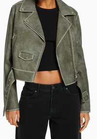 Zelená faux kůže zkrácená motorkářská bunda s klopovým límcem, předním zipem a detaily na cvakání. V kombinaci s černým žebrovaným topem a černými džíny.