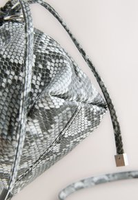 Sac en cuir synthétique imitation peau de serpent gris et blanc avec une surface texturée, doté d'une bandoulière tressée et d'un accent métallique, forme angulaire.