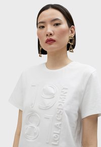 Camiseta de algodón blanca con el número 1981 elevado y texto de marca, cuello redondo, mangas cortas y una textura suave.