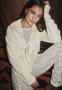Gekürzter cremefarbener Cardigan mit breitem Kragen und Knopfleiste, kombiniert mit enganliegenden Spitzenhosen in Creme. Strukturiertes Material mit gerippten Ärmeln.