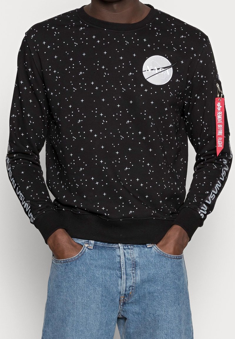 Svart sweatshirt med mönster av vita stjärnor, NASA-logotyp på bröstet, "NASA"-text på ärmarna och röd etikett "Remove Before Flight" på vänster arm.