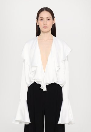 Valkoinen röyhelöblouse syvällä V-kaula-aukolla ja pitkillä hihoilla. Kangas on sileä. Yhdistetty mustiin korkea vyötäröisiin housuihin.