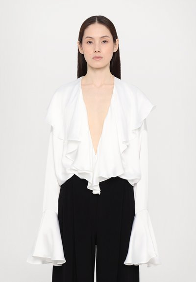 Witte gerimpelde blouse met een diepe V-hals en lange mouwen. De stof heeft een gladde afwerking. Gecombineerd met zwarte hooggerokte broeken.