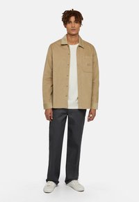 Tan corduroy overhemd met knoopsluiting en borstzak, gecombineerd met donkergrijze broek en witte sneakers, met een eenvoudig ontwerp.