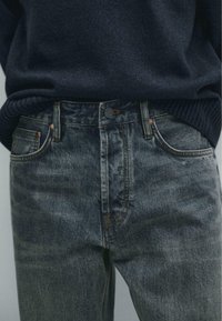 Dark blue denim jeans with a slight fade, five-pocket design, metal buttons, and contrasting orange stitching. Textured fabric with minimal distressing.