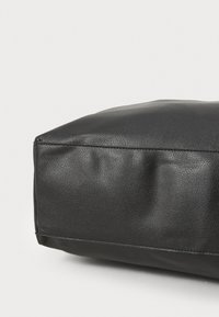 Črna usnjena torba z teksturirano površino, kvadratne oblike in subtilno šivano podrobnostjo na strani, ki prikazuje gladko končno obdelavo.