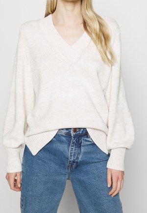 Woman wearing a white V-neck sweater with ribbed cuffs and hem, paired with high-waisted blue jeans.