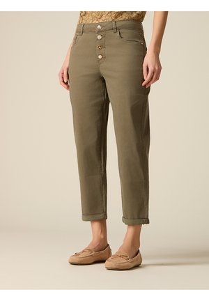 Kobieta w oliwkowozielonych, krótkich spodniach z czterema guzikami z przodu oraz beżowych mokasynach, stojąca na jednolitym tle.