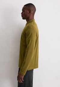 Langarm-Poloshirt in Olivgrün, mit klassischem Kragen, schmaler Passform und gerippten Bündchen. Kombiniert mit dunklen Jeans.