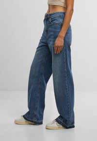 Wide-leg blue denim jeans with a faded wash, high waist, and raw hems, paired with white sneakers.