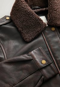 Brun læderjakke med en brun shearlingkrave, udstyret med en klaplomme med en messingstuds og tekstureret materiale.