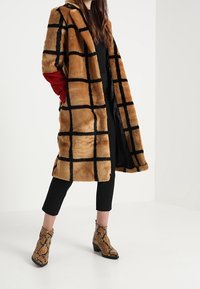 Abrigo de peluche sintético de cuadros marrones con detalles rojos, abertura frontal y patrón de cuadrícula negra. Combinado con pantalones negros y botines de tobillo con estampado de serpiente.