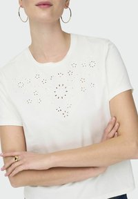 Hvid bomulds T-shirt med blomstret øjeletsdetalje på fronten, korte ærmer og rund halsudskæring. Glat tekstur, afslappet pasform.