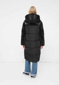 Zwarte lange pufferjas met een glanzende afwerking, capuchon en horizontale naden, gecombineerd met lichtblauwe flared jeans en zwarte sneakers.