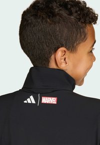 Zwarte jas met hoge kraag, voorzien van een wit Adidas-logo en een rood Marvel-logo op de achterkant, gemaakt van glad en rekbaar materiaal.