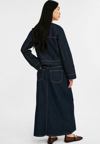 Donkerblauwe spijkerjacks en maxi-jurk set met lange mouwen, zichtbare stiksels en twee achterzakken. Platte schoenen met een patroon.