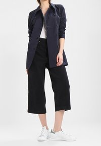 Námořnický trenčkot s přiléhavým střihem, s knoflíkovými detaily, v kombinaci s černými zkrácenými kalhotami a bílými teniskami s zelenými akcenty.