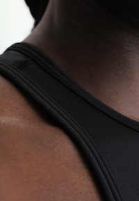 Κοντινό μιας ώμου και λαιμού που φορά μαύρο αμάνικο με ορατές ραφές και λεία σκούρα υφή δέρματος.