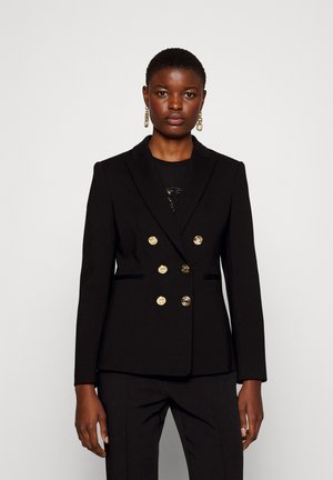 Blazer nero doppiopetto con bottoni dorati, vestibilità sartoriale, rever a punte e cuciture frontali. Indossato sopra una maglietta nera.