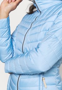 Světle modrá péřová bunda s prošívanou strukturou, vysokým límcem a zapínáním na zip. Obsahuje boční kapsu se zlatým jezdcem.