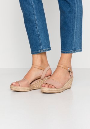 Fødder iført lysebrune kilehælssandaler med ankelremme og blå jeans, der er croppede over anklerne, på hvidt gulv.
