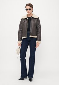 Kvinne iført svart saueskinnsjakke, mørkeblå jeans, svarte spisse hæler, solbriller, holder en liten hvit håndveske mot hvit bakgrunn.