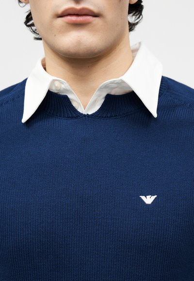 Νεαρός άνδρας που φοράει λευκό πουκάμισο με γιακά κάτω από μπλε σκούρο πλεκτό πουλόβερ με μικρό λευκό κεντητό λογότυπο στο στήθος.