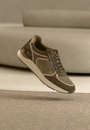 Baskets vert olive avec garniture beige, fermeture éclair latérale et lacets, flottant au-dessus d'un sol recouvert de moquette beige et d'un canapé beige clair en arrière-plan.