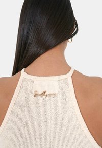 Cremefärgad stickad halterneck-top med en texturerad yta och en logotyp i guldton på baksidan. Remmarna är släta och rundade.