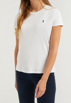 Nainen pukeutuneena yksinkertaiseen valkoiseen lyhythihaiseen t-paitaan, jossa on pieni musta golfarin logo rinnassa, yhdistettynä tummansinisiin housuihin.