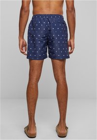 Shorts de bain bleu marine avec des motifs blancs de têtes de mort, de bateaux et d'ancres croisées. Taille élastique avec cordon de serrage et poche arrière.