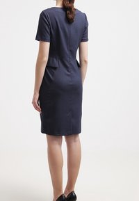 Kvinna som bär en figursydd, kortärmad marinblå klänning med fickdetaljer vid midjan, stående med ryggen mot kameran på en enfärgad bakgrund.