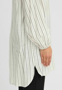 Camicia bianca a maniche lunghe con strisce verticali scure, con polsini elasticizzati e orlo curvo, mostrata accanto a un indumento nero.