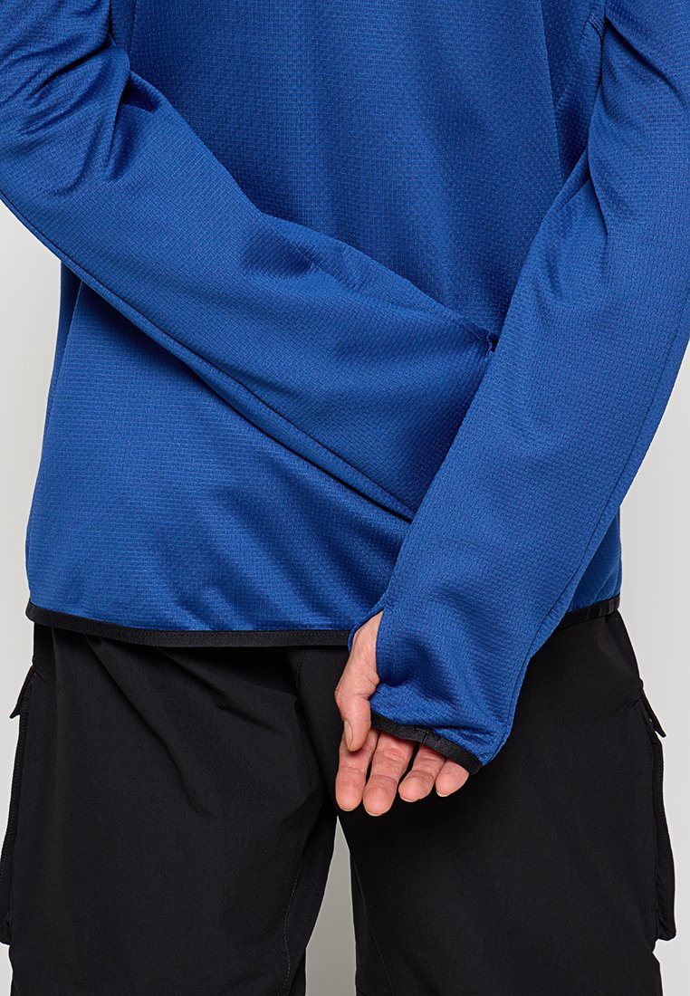 Chemise bleue à manches longues en tissu texturé, avec une coupe ajustée et un ourlet noir contrastant. Main visible glissée dans la manche.