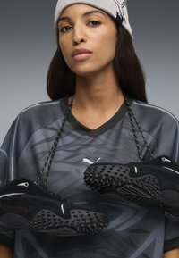 Scarpe da atletica nere con suole testurizzate, sospese da lacci neri, su uno sfondo di una maglia grafica grigia con il logo Puma.