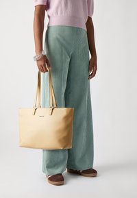 Borsa a tote gialla in pelle con doppi manici, finitura liscia, logo sulla parte anteriore, abbinata a pantaloni in corduroy verde chiaro e top in maglia rosa.
